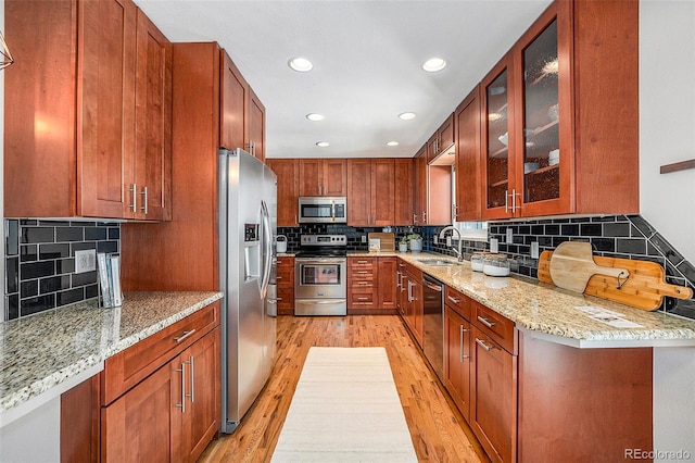 kitchen with light stone counters, a sink, appliances with stainless steel finishes, light wood finished floors, and glass insert cabinets