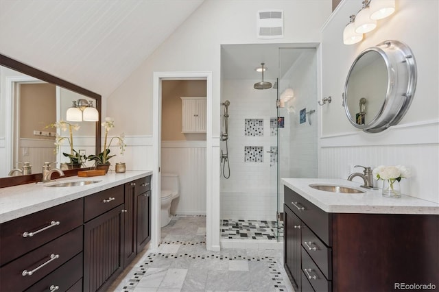 bathroom featuring a stall shower, lofted ceiling, toilet, wainscoting, and two vanities