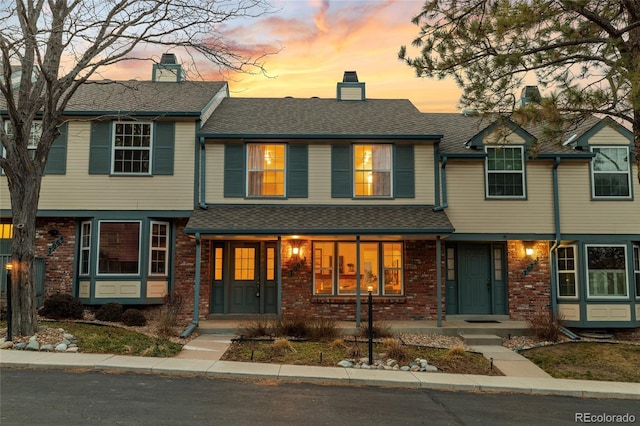 townhome / multi-family property with covered porch, brick siding, roof with shingles, and a chimney