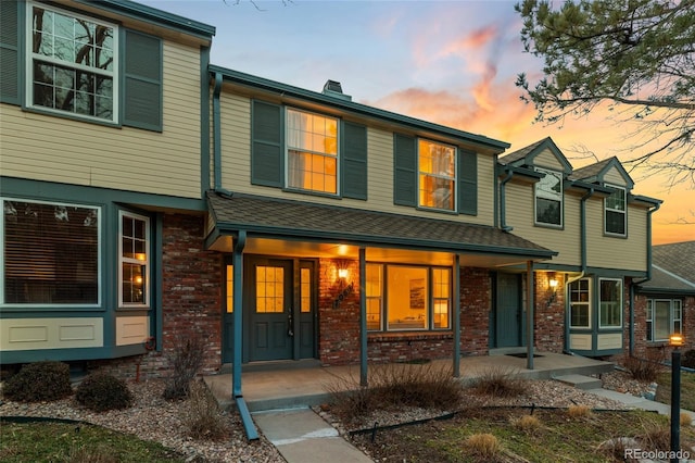 multi unit property with brick siding, covered porch, a chimney, and a shingled roof