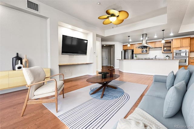 living room featuring light wood-type flooring, visible vents, a ceiling fan, baseboards, and a raised ceiling