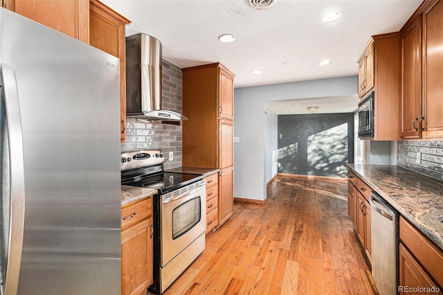 kitchen featuring appliances with stainless steel finishes, backsplash, light hardwood / wood-style floors, dark stone counters, and wall chimney exhaust hood