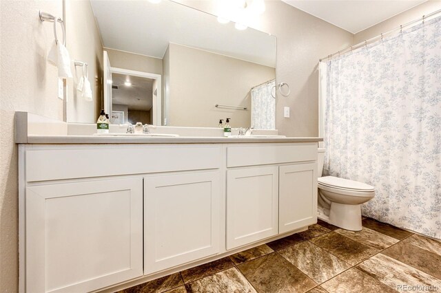 full bath with double vanity, curtained shower, toilet, stone finish floor, and a sink