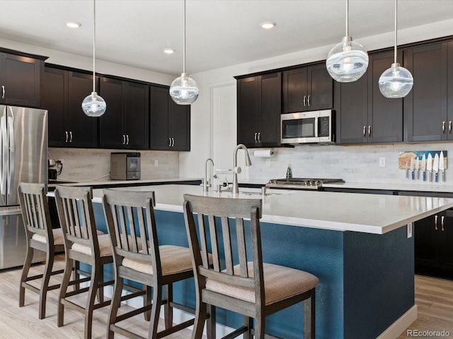 kitchen with light wood finished floors, tasteful backsplash, pendant lighting, an island with sink, and appliances with stainless steel finishes