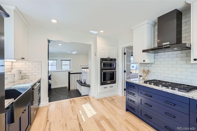 kitchen featuring wall chimney exhaust hood, multiple ovens, light wood-style floors, stainless steel gas stovetop, and white cabinetry