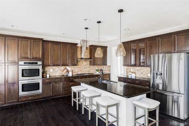 kitchen with wall chimney exhaust hood, sink, dark stone countertops, appliances with stainless steel finishes, and an island with sink