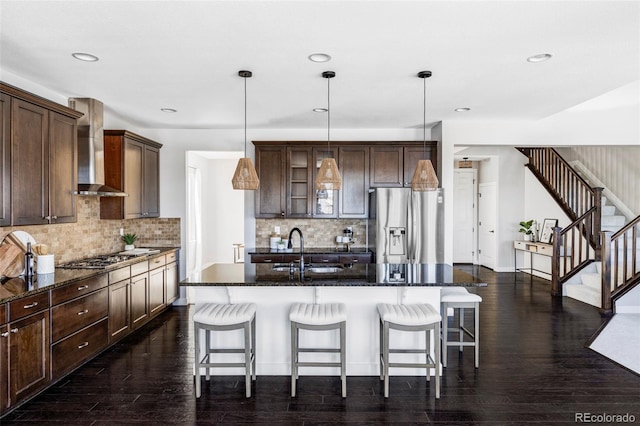 kitchen featuring dark brown cabinetry, pendant lighting, stainless steel appliances, a kitchen island with sink, and wall chimney range hood