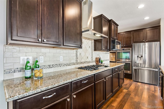 kitchen featuring wall chimney range hood, light stone countertops, dark brown cabinetry, dark wood finished floors, and appliances with stainless steel finishes