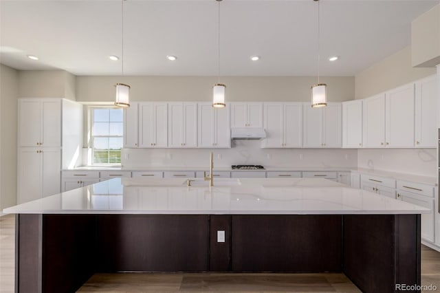56159 E Tomahawk Ave, Strasburg, CO 80136 - kitchen with under cabinet range hood, stainless steel gas stovetop, a large island with sink, light wood finished floors, and recessed lighting