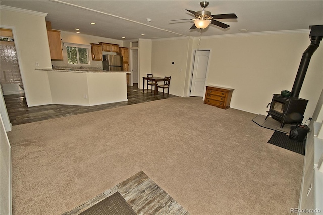 unfurnished living room with ceiling fan, a wood stove, crown molding, and dark colored carpet