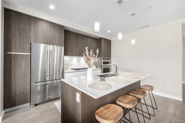 kitchen featuring a breakfast bar, decorative light fixtures, light wood-type flooring, stainless steel appliances, and a kitchen island with sink