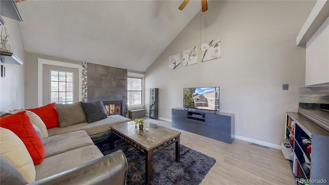 living area featuring baseboards, ceiling fan, a tile fireplace, light wood-style floors, and high vaulted ceiling