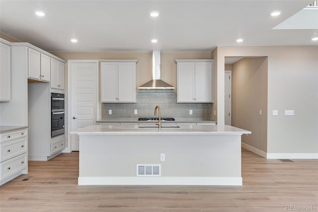 kitchen featuring an island with sink, white cabinets, light stone counters, and wall chimney exhaust hood