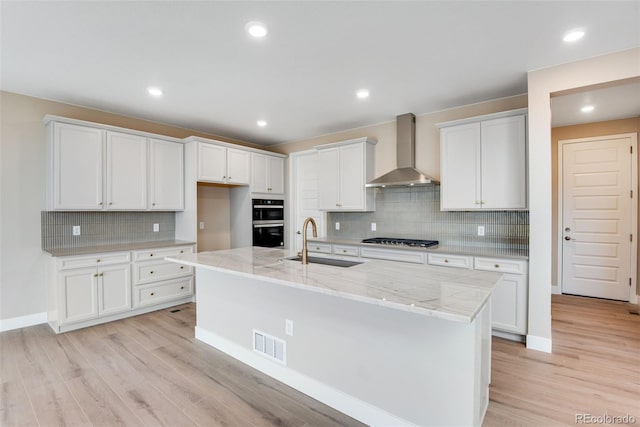 kitchen featuring sink, a center island with sink, wall chimney range hood, stainless steel gas stovetop, and white cabinets