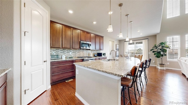 kitchen featuring appliances with stainless steel finishes, light stone counters, a kitchen island with sink, sink, and pendant lighting