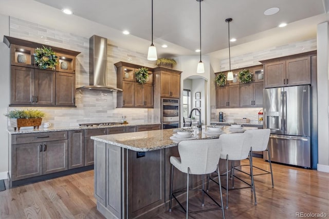 kitchen featuring wall chimney range hood, a center island with sink, appliances with stainless steel finishes, wood finished floors, and arched walkways