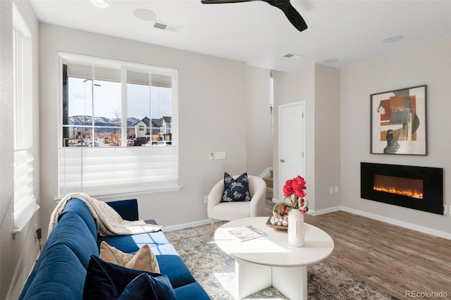 living area with wood finished floors, a ceiling fan, visible vents, baseboards, and a glass covered fireplace