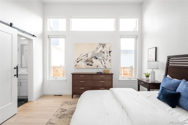 bedroom with baseboards, light wood finished floors, ensuite bathroom, a towering ceiling, and a barn door