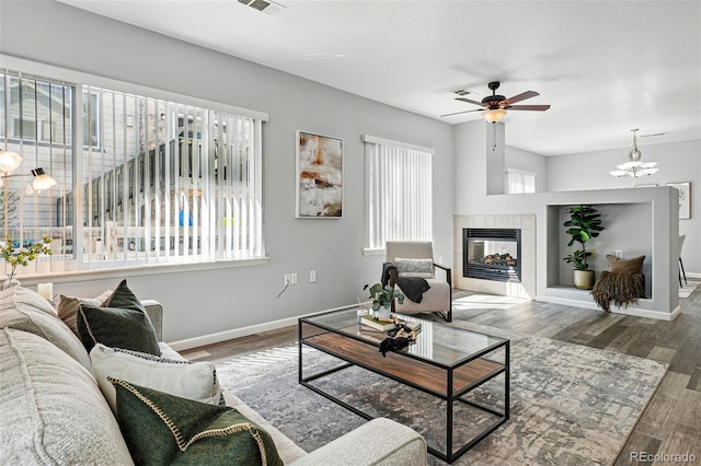 living room featuring wood finished floors, a tiled fireplace, baseboards, and ceiling fan with notable chandelier