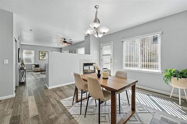 dining room featuring wood finished floors, a tiled fireplace, baseboards, and ceiling fan with notable chandelier
