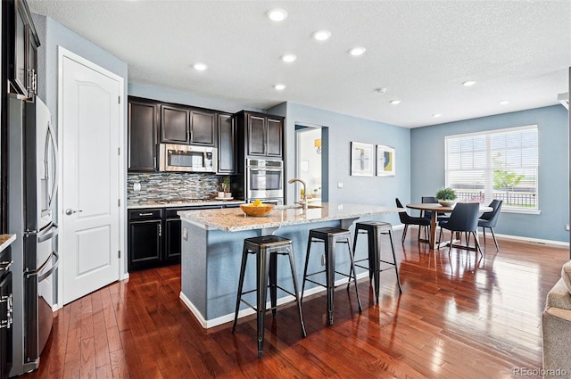 7426 E 148th Pl, Thornton, CO 80602 - kitchen featuring stainless steel appliances, dark wood finished floors, backsplash, light stone counters, and a center island with sink