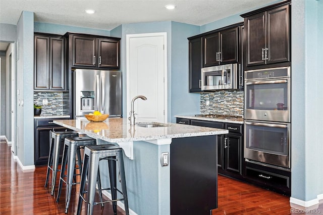 7426 E 148th Pl, Thornton, CO 80602 - kitchen with appliances with stainless steel finishes, a sink, dark wood finished floors, light stone counters, and a center island with sink