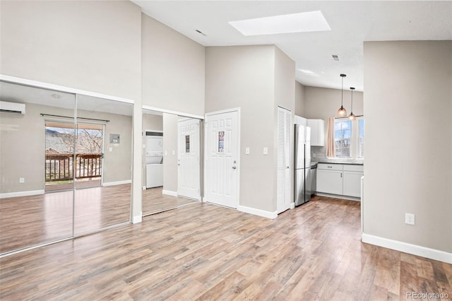 unfurnished living room featuring a wall unit AC, a skylight, a towering ceiling, light wood-style flooring, and baseboards