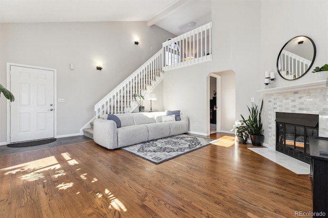 living area featuring beam ceiling, wood finished floors, baseboards, a brick fireplace, and stairs