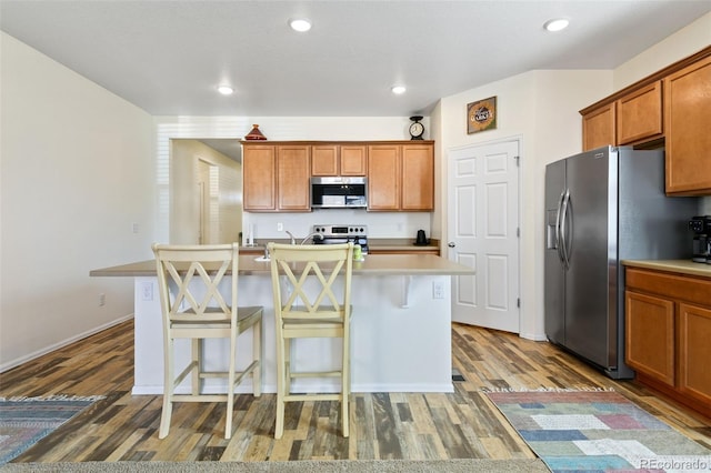 kitchen featuring a kitchen island with sink, a kitchen breakfast bar, wood finished floors, appliances with stainless steel finishes, and light countertops