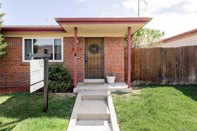 view of exterior entry with a lawn, brick siding, and fence