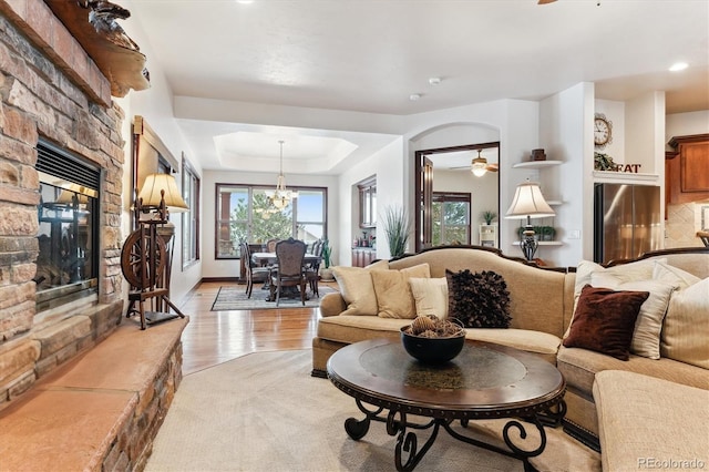 living room featuring a tray ceiling, ceiling fan with notable chandelier, a fireplace, and light hardwood / wood-style floors