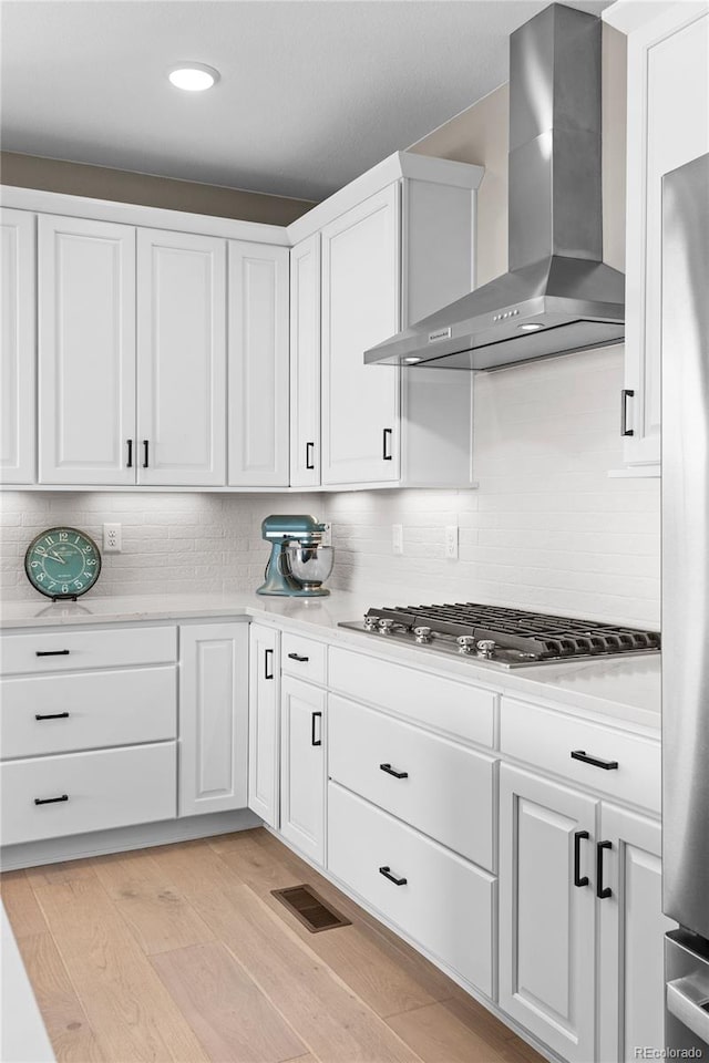 kitchen with decorative backsplash, appliances with stainless steel finishes, wall chimney exhaust hood, light hardwood / wood-style floors, and white cabinetry