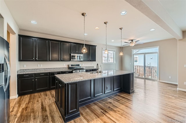 kitchen with light stone countertops, decorative light fixtures, an island with sink, appliances with stainless steel finishes, and ceiling fan