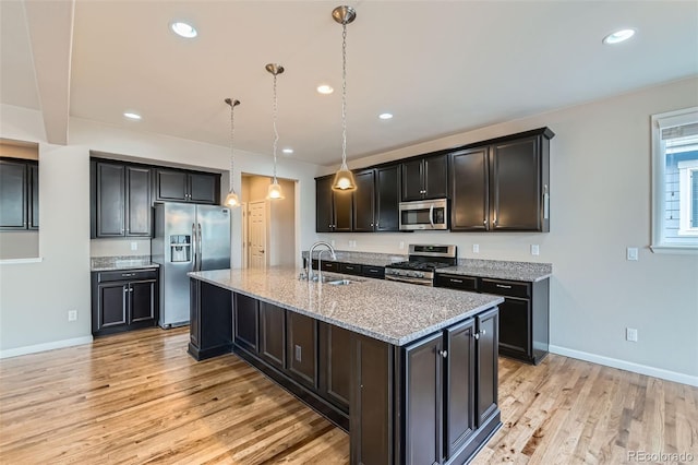 kitchen with light stone countertops, light hardwood / wood-style flooring, pendant lighting, an island with sink, and appliances with stainless steel finishes