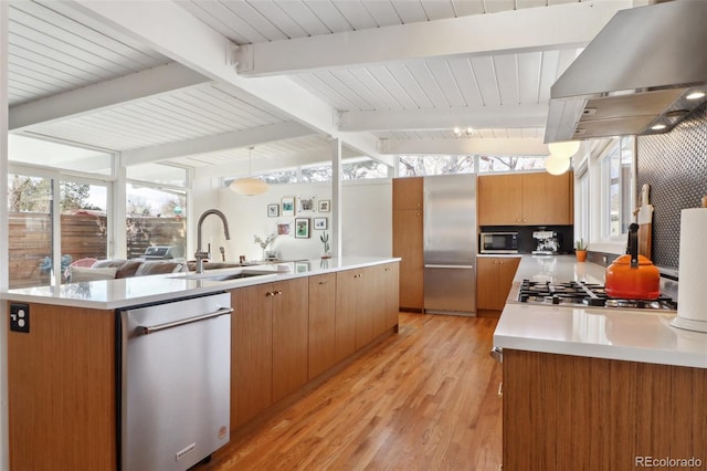 kitchen with vaulted ceiling with beams, brown cabinetry, stainless steel appliances, wall chimney exhaust hood, and a sink