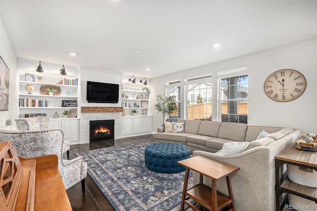 living area featuring recessed lighting, built in features, dark wood-type flooring, and a warm lit fireplace