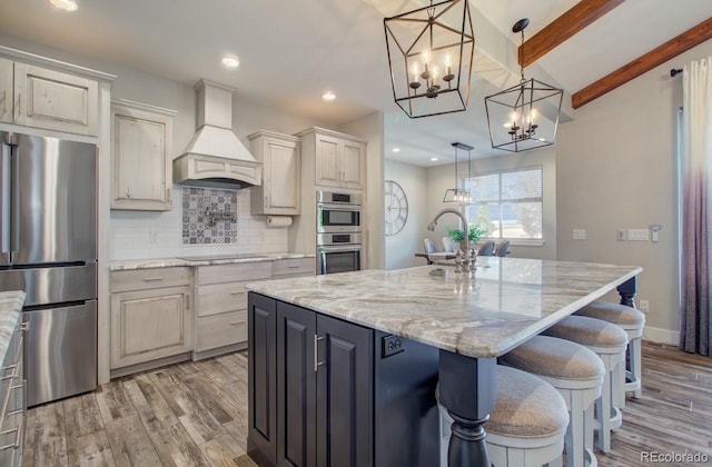 kitchen with appliances with stainless steel finishes, light hardwood / wood-style flooring, premium range hood, and an island with sink