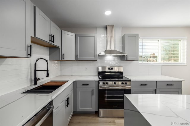 kitchen with stainless steel appliances, gray cabinetry, wall chimney exhaust hood, light stone counters, and sink