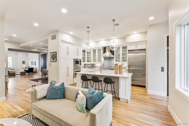 kitchen featuring a kitchen island with sink, white cabinetry, built in appliances, decorative light fixtures, and wall chimney exhaust hood