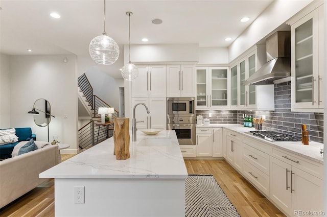 kitchen featuring decorative light fixtures, white cabinets, a kitchen island with sink, stainless steel appliances, and wall chimney range hood