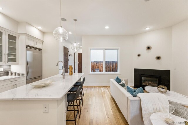 kitchen featuring stainless steel built in refrigerator, light stone counters, hanging light fixtures, an island with sink, and white cabinets