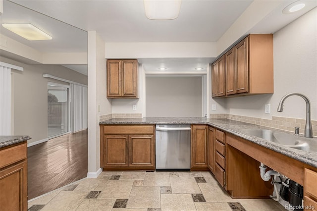 kitchen with stone tile floors, stainless steel dishwasher, brown cabinetry, a sink, and baseboards