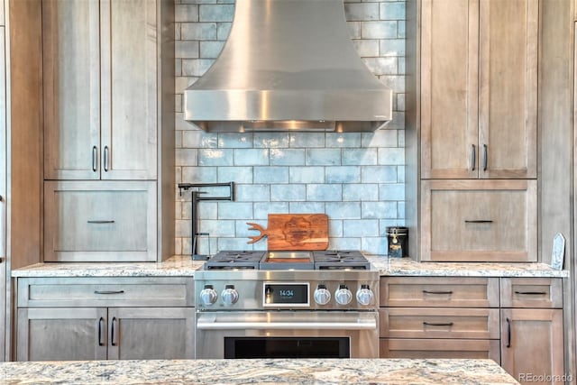 kitchen featuring decorative backsplash, light stone counters, wall chimney exhaust hood, and high end stainless steel range oven