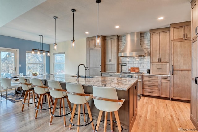 kitchen featuring wall chimney exhaust hood, a large island with sink, pendant lighting, high quality appliances, and light wood-type flooring