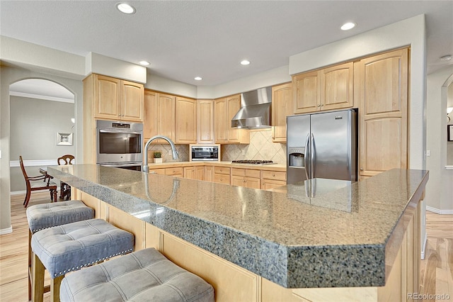 14878 E Aberdeen Ave, Centennial, CO 80016 - kitchen featuring appliances with stainless steel finishes, arched walkways, wall chimney range hood, light brown cabinetry, and recessed lighting