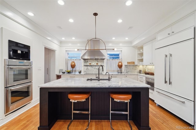 kitchen with stainless steel double oven, white cabinets, sink, paneled refrigerator, and a center island with sink