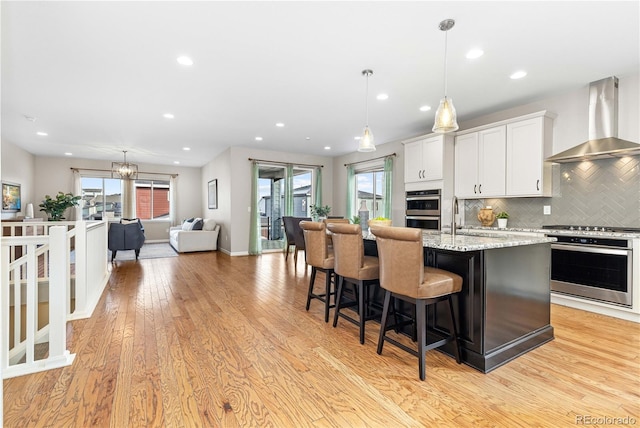 kitchen featuring light stone countertops, an island with sink, white cabinets, decorative light fixtures, and wall chimney exhaust hood