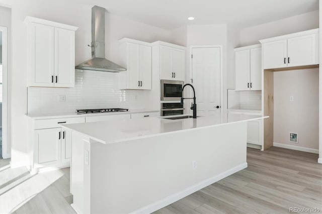 kitchen featuring wall chimney exhaust hood, stainless steel appliances, a kitchen island with sink, white cabinets, and light hardwood / wood-style floors