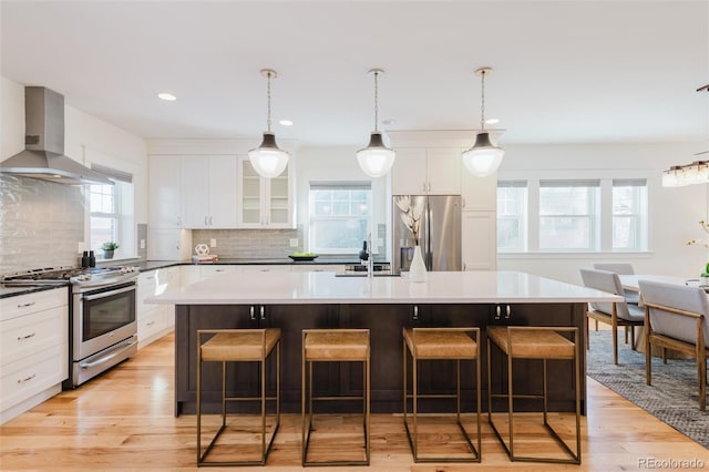 kitchen with a kitchen island with sink, wall chimney exhaust hood, appliances with stainless steel finishes, tasteful backsplash, and decorative light fixtures