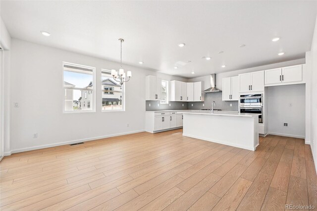 kitchen with a sink, a chandelier, light wood-style flooring, wall chimney exhaust hood, and white cabinetry